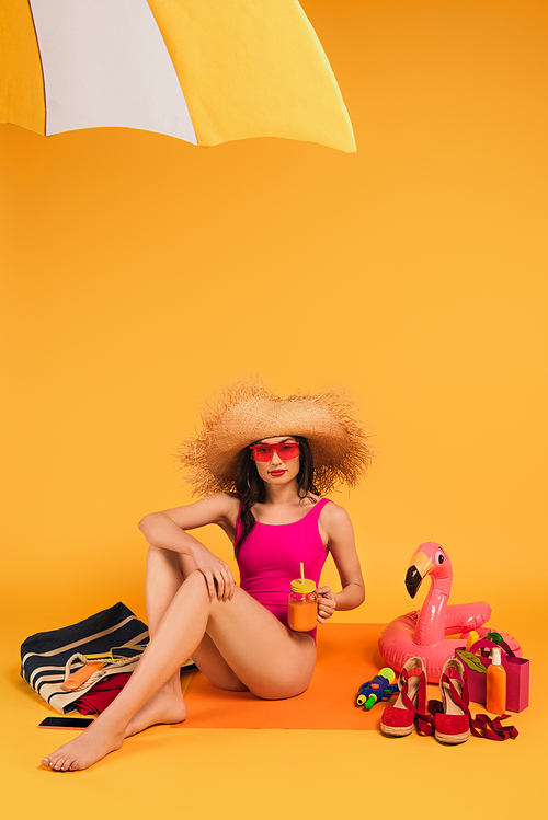 stylish woman in straw hat, sunglasses and pink swimsuit holding glass with orange juice near water gun and inflatable ring on yellow
