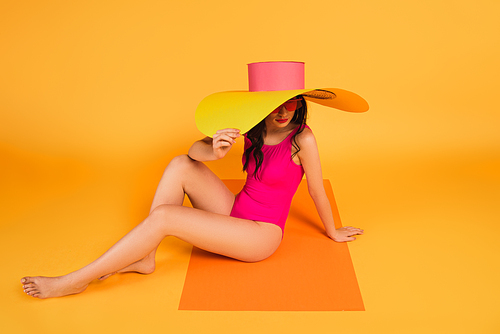 stylish girl in straw hat and bathing suit covering face while sitting on yellow