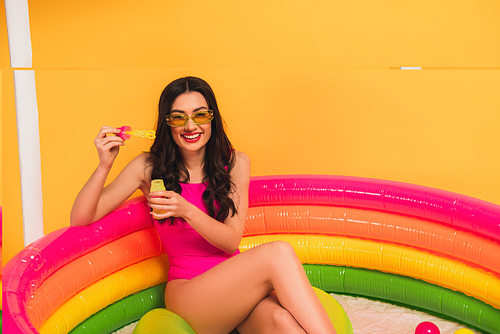 happy woman in swimsuit holding bottle with soap bubbles while sitting in inflatable pool on yellow