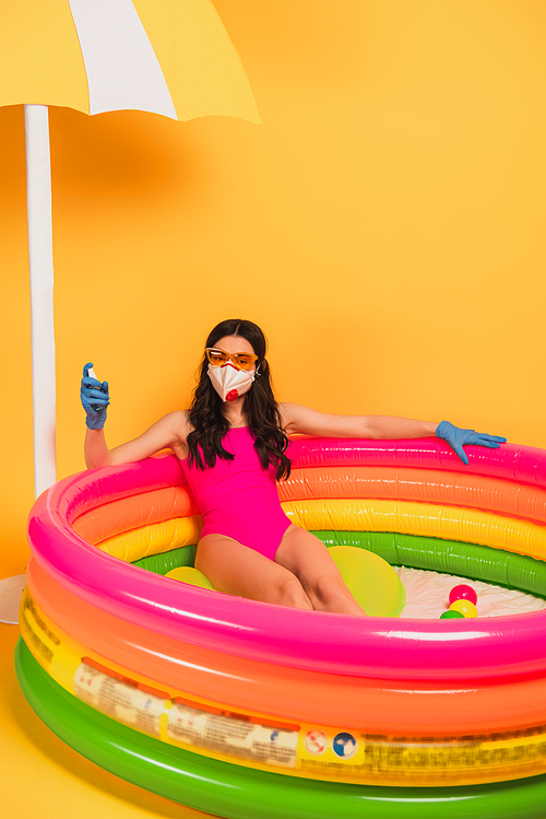 young woman in bathing suit, sunglasses, latex gloves and medical mask sitting in inflatable pool and holding sanitizer on yellow