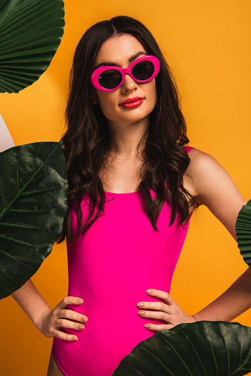 stylish girl in sunglasses and swimsuit standing with hands on hips near green palm leaves on yellow