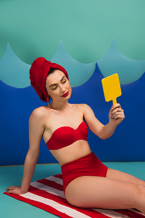 beautiful woman in red towel and swimsuit looking at mirror near blue paper cut waves