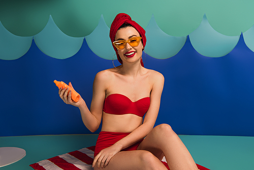happy woman in red swimsuit and sunglasses holding sunscreen near paper cut waves
