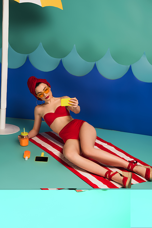 attractive woman in swimsuit and sunglasses taking selfie near paper cut waves