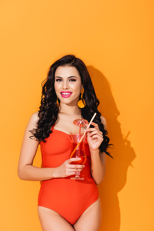 cheerful woman in swimsuit holding glass with cocktail and standing on orange