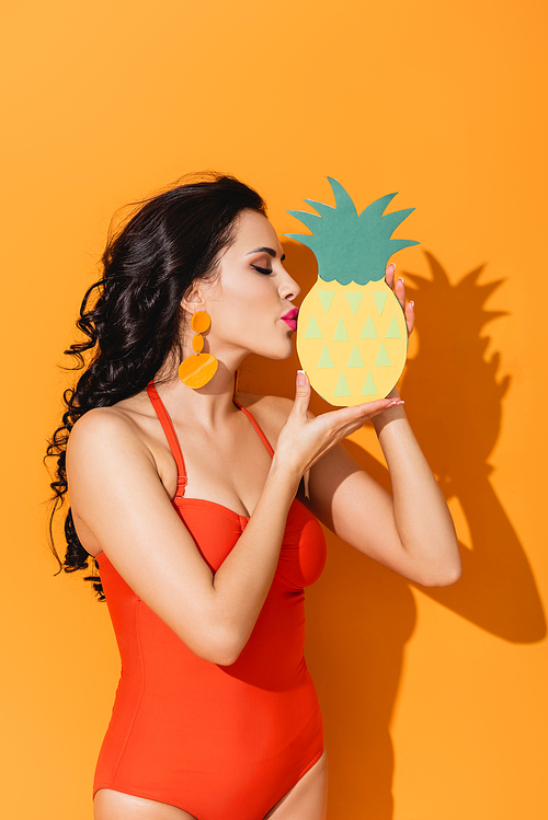 side view of attractive woman in swimsuit kissing paper cut pineapple on orange