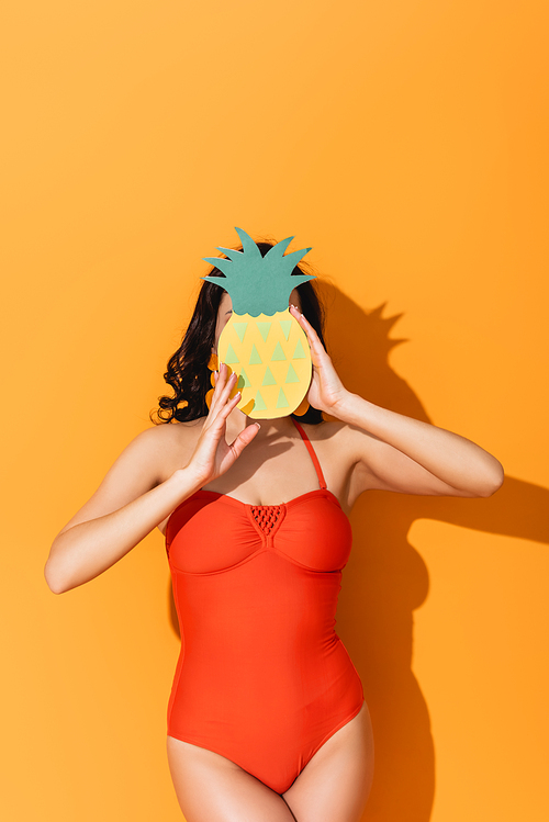 young woman in swimsuit covering face with paper cut pineapple on orange