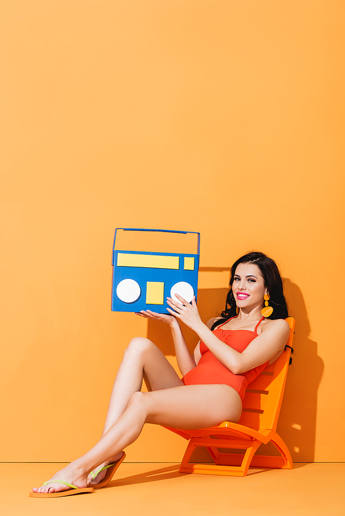 happy woman in swimsuit holding paper boombox while sitting on deck chair on orange