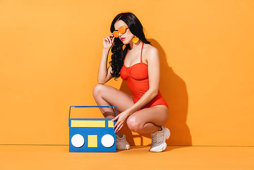 stylish woman in sneakers and bathing suit touching sunglasses while sitting near paper cut boombox on orange