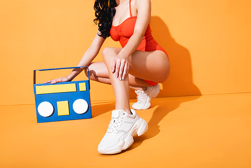 cropped view of stylish woman in sneakers and bathing suit sitting near paper cut boombox on orange