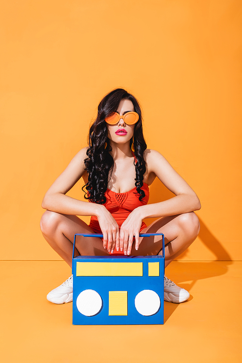 stylish girl in sneakers, bathing suit and sunglasses sitting near paper cut boombox on orange