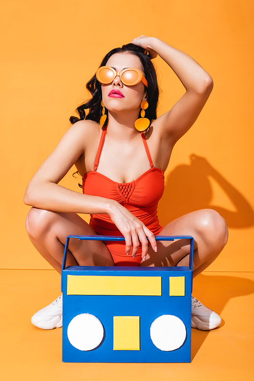 stylish girl in sneakers, bathing suit and sunglasses sitting near paper cut boombox and touching hair on orange