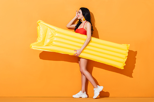 side view of cheerful girl in swimsuit and sneakers holding inflatable mattress on orange