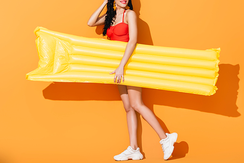 cropped view of happy girl in swimsuit and sneakers holding inflatable mattress on orange