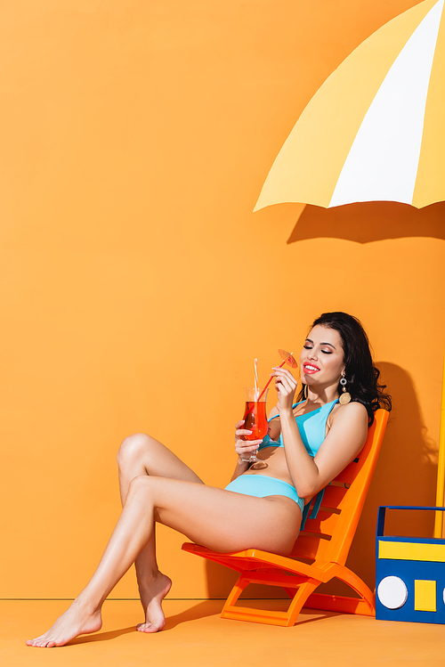 smiling woman in swimsuit sitting on deck chair near paper boombox and umbrella while looking at cocktail on orange