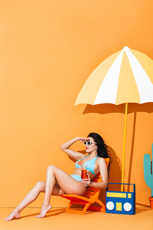 stylish woman in sunglasses and swimsuit sitting on deck chair near paper boombox and umbrella while holding cocktail on orange