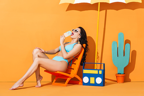 trendy woman in sunglasses and swimwear sitting on deck chair near boombox and umbrella while licking paper ice cream on orange