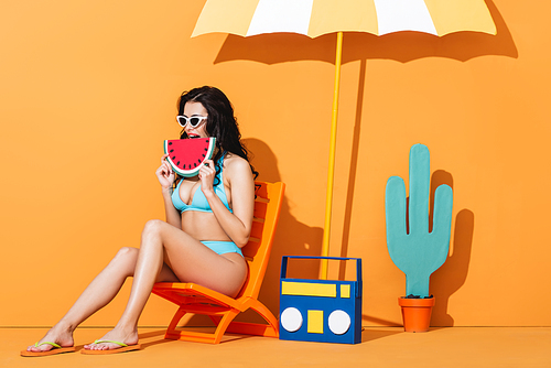 young woman in sunglasses and swimsuit sitting on deck chair near boombox and umbrella while holding paper watermelon on orange