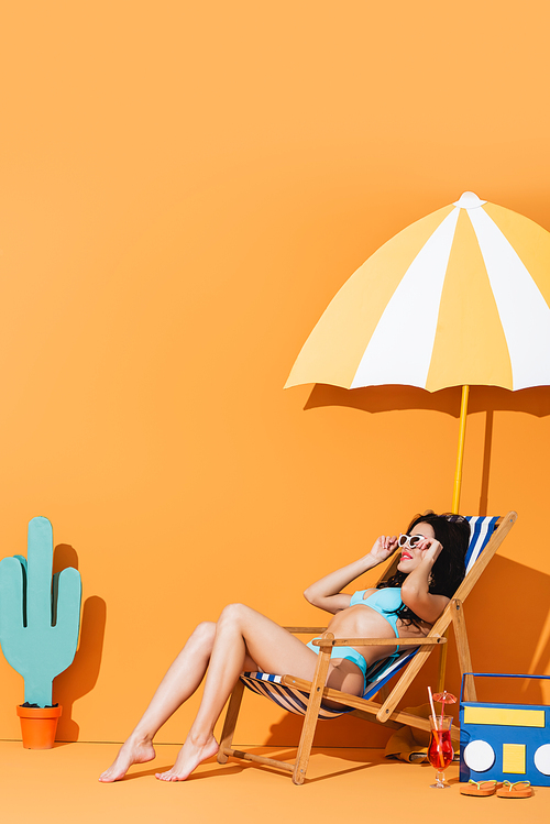 trendy woman in sunglasses and swimwear sitting on deck chair near cocktail, flip flops, paper boombox, cactus and umbrella on orange