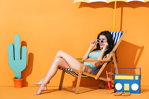 happy woman in swimwear touching sunglasses and sitting on deck chair near cocktail, flip flops, paper boombox and cactus on orange