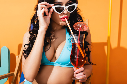 young woman in swimwear touching sunglasses and drinking cocktail on orange