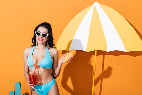 happy woman in swimwear and sunglasses holding sunscreen and cocktail near paper umbrella on orange