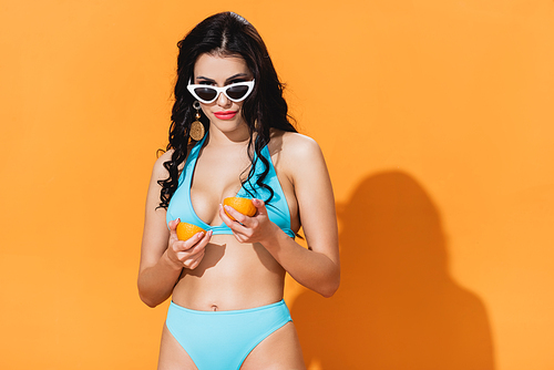 stylish woman in bathing suit and sunglasses holding organic fruit halves on orange