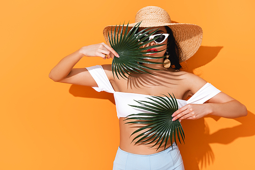fashionable girl in swimsuit, sunglasses and straw hat holding palm leaves on orange