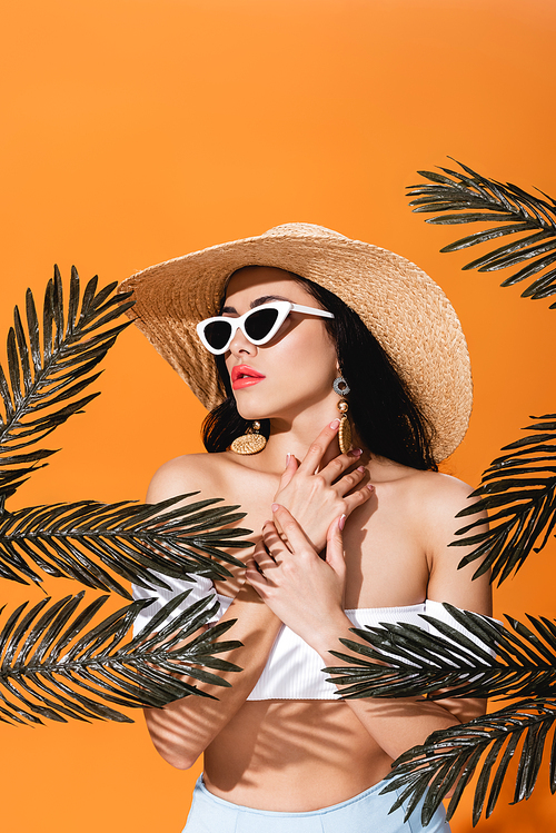 stylish woman in sunglasses and straw hat touching neck near palm leaves isolated on orange