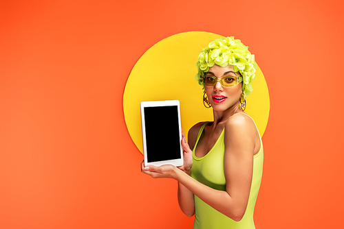 Girl in hat with decorative flowers  and presenting digital tablet with yellow circle behind on orange