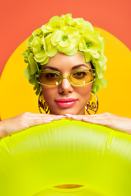 Portrait of girl in hat with decorative flowers and sunglasses smiling and putting hands on swim ring on yellow and orange