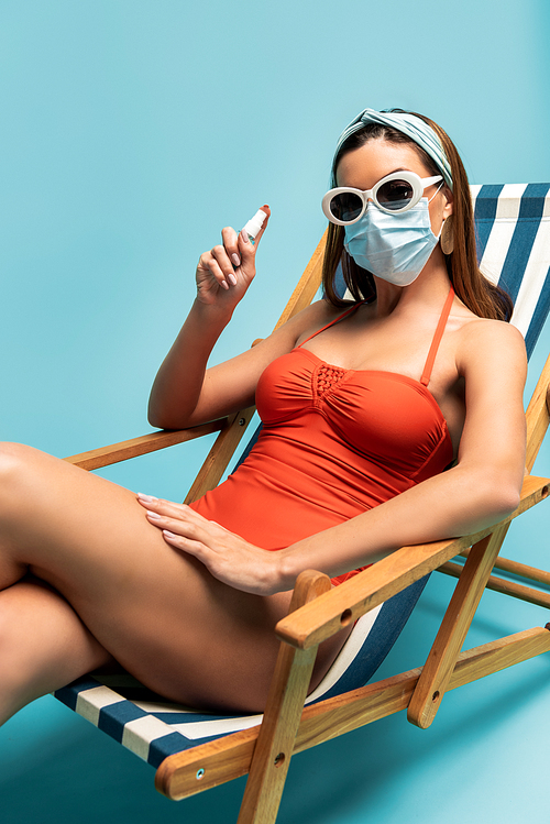 Woman in sunglasses and medical mask showing bottle of hand sanitizer on deckchair on blue