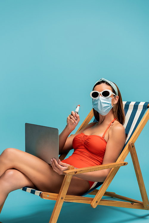 Freelancer in medical mask with laptop showing bottle of hand sanitizer on deckchair on blue