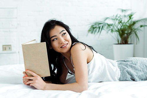 positive asian girl holding book while lying on bed at home