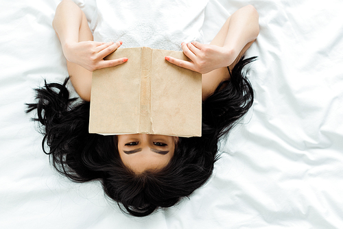 top view of asian girl covering face with book while lying on bed