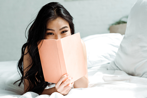 asian young woman covering face with book while lying on bed