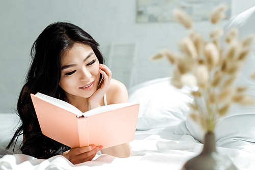 selective focus of positive asian girl reading book while lying on bed at home