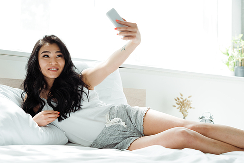 selective focus of cheerful brunette asian woman taking selfie in bedroom