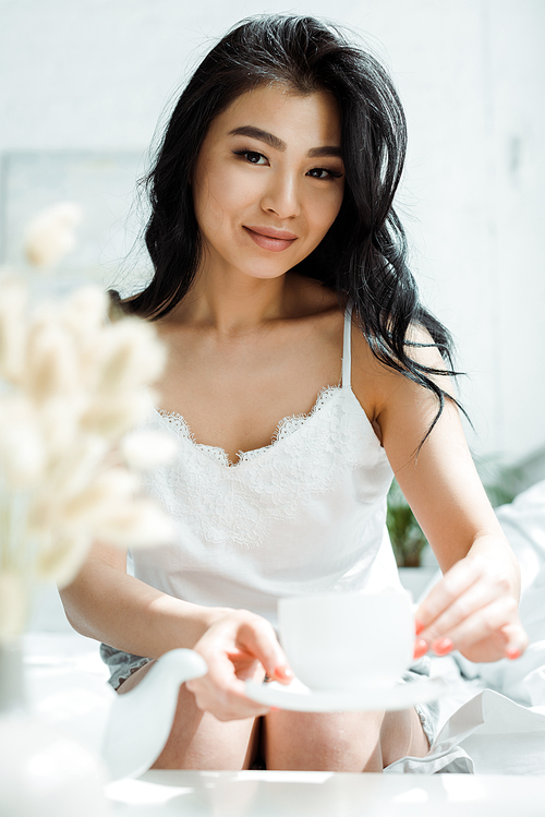 selective focus of happy brunette thai woman holding saucer and cup of tea