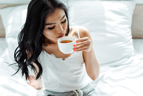 selective focus of brunette thai woman holding cup with tea while sitting in bedroom