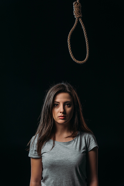 depressed young woman looking away while standing under hanging noose isolated on balck