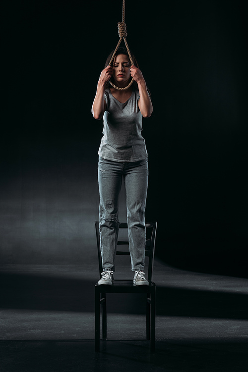 depressed young woman committing suicide while standing on chair and putting noose on neck on black background