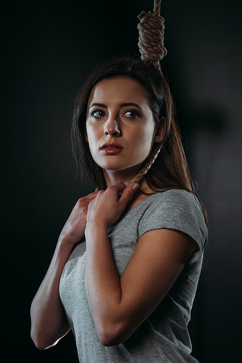 frustrated young woman looking away while standing with noose on neck on black background