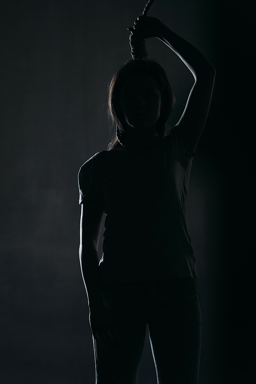 depressed woman committing suicide while standing with noose on neck in darkness on black background