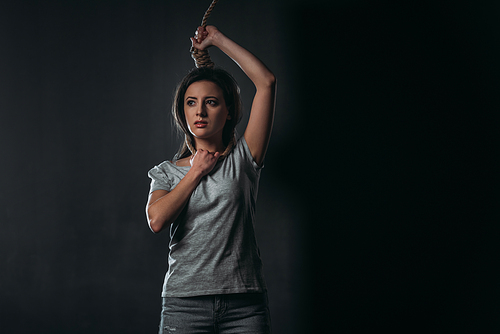 despaired woman looking away while standing with noose on neck on black background