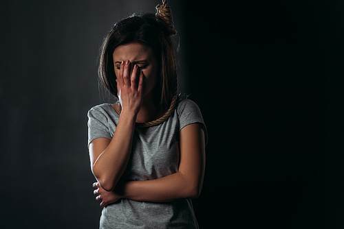 crying woman covering face with hand while standing with noose on neck on black background