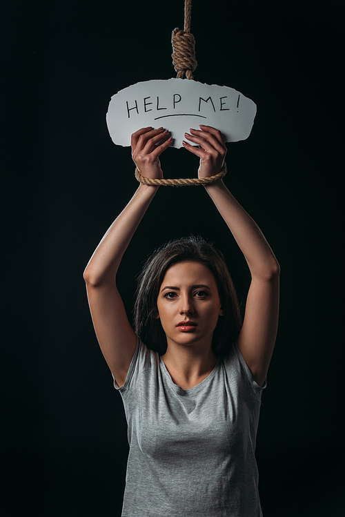 frustrated young woman holding paper with help me lettering while standing under hanging noose and  isolated on black