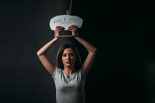 frustrated young woman holding paper with help me inscription while standing under hanging noose and  on black background
