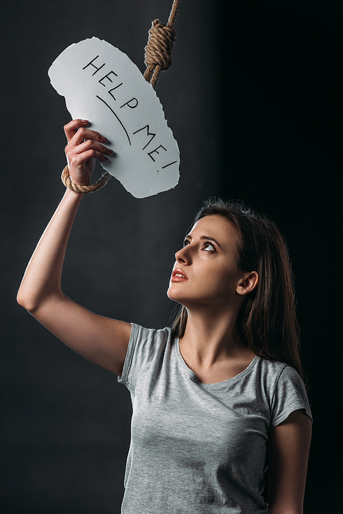 depressed young woman holding paper with help me lettering near hanging noose on black background