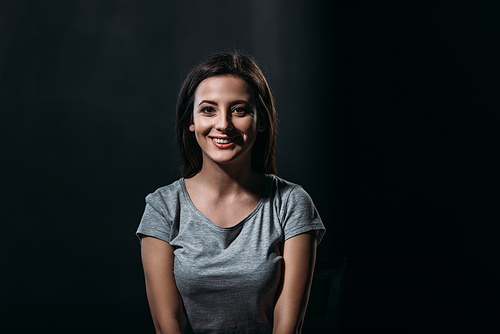 happy young woman smiling at camera while sitting in darkness isolated on black